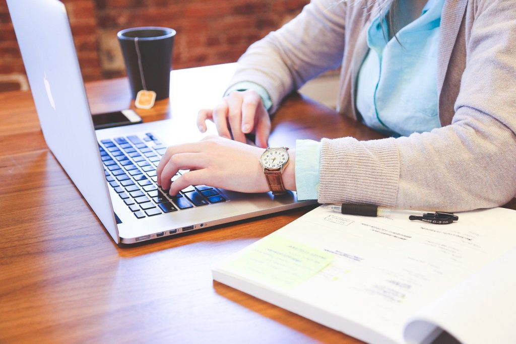Une personne avec une chemise et un cardigan écrit sur son ordinateur portable avec un carnet ouvert et une tasse à côté.