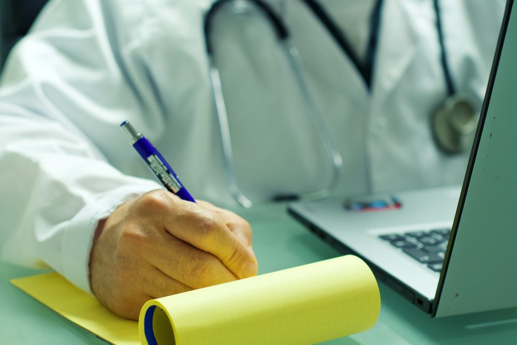 Une personne en blouse blanche avec un stéthoscope écrit avec un stylo dans un carnet jaune.