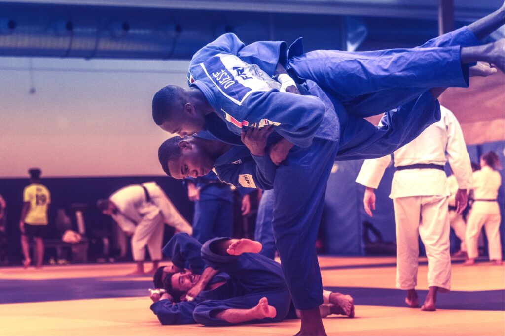 Deux judokas en kimonos bleus s'entraînent sur un tatami avec d'autres judokas en kimonos blancs autour.