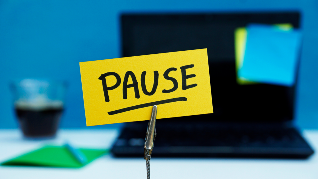 Un papier avec écrit "PAUSE" est affiché devant un ordinateur portable, une pochette et un verre rempli de café.