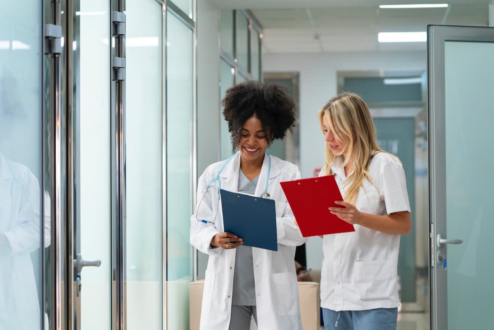 Deux femmes en blouses blanches se parlent dans un couloir d'hôpital et tiennent des pochettes.