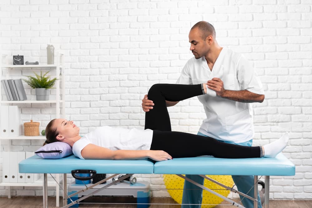 Une patiente est allongée sur une table de kinésithérapeute, un homme en blouse blanche lui tient la jambe et le pied.
