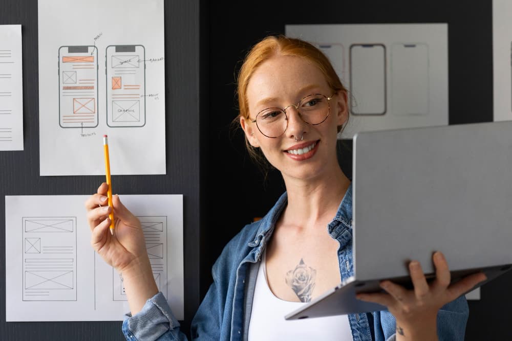 Une femme tiens un ordinateur et pointe avec un stylo une interface web pour téléphone sur une feuille.